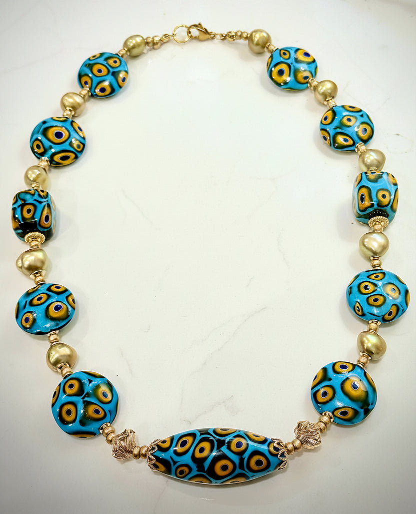 A handmade glass bead necklace featuring vibrant turquoise blue beads with yellow and black concentric eye patterns, interspersed with small 12k gold filled twisted lantern beads and a larger matching blue bead at the center, displayed on a white background.